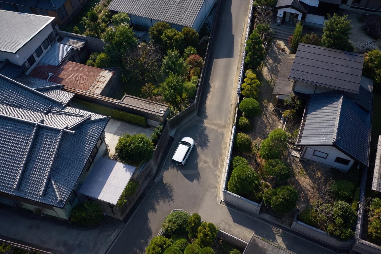 住宅街の細い道の角を曲がる白いSUVをドローンで見下ろした写真